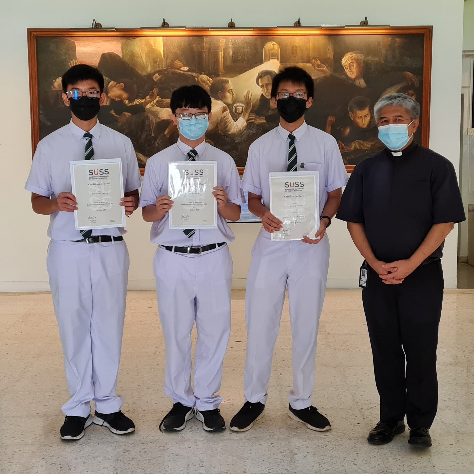 Three masked students and a priest hold SUSS Certificate of Merit awards for the Business Plan Competition, before a large mural.